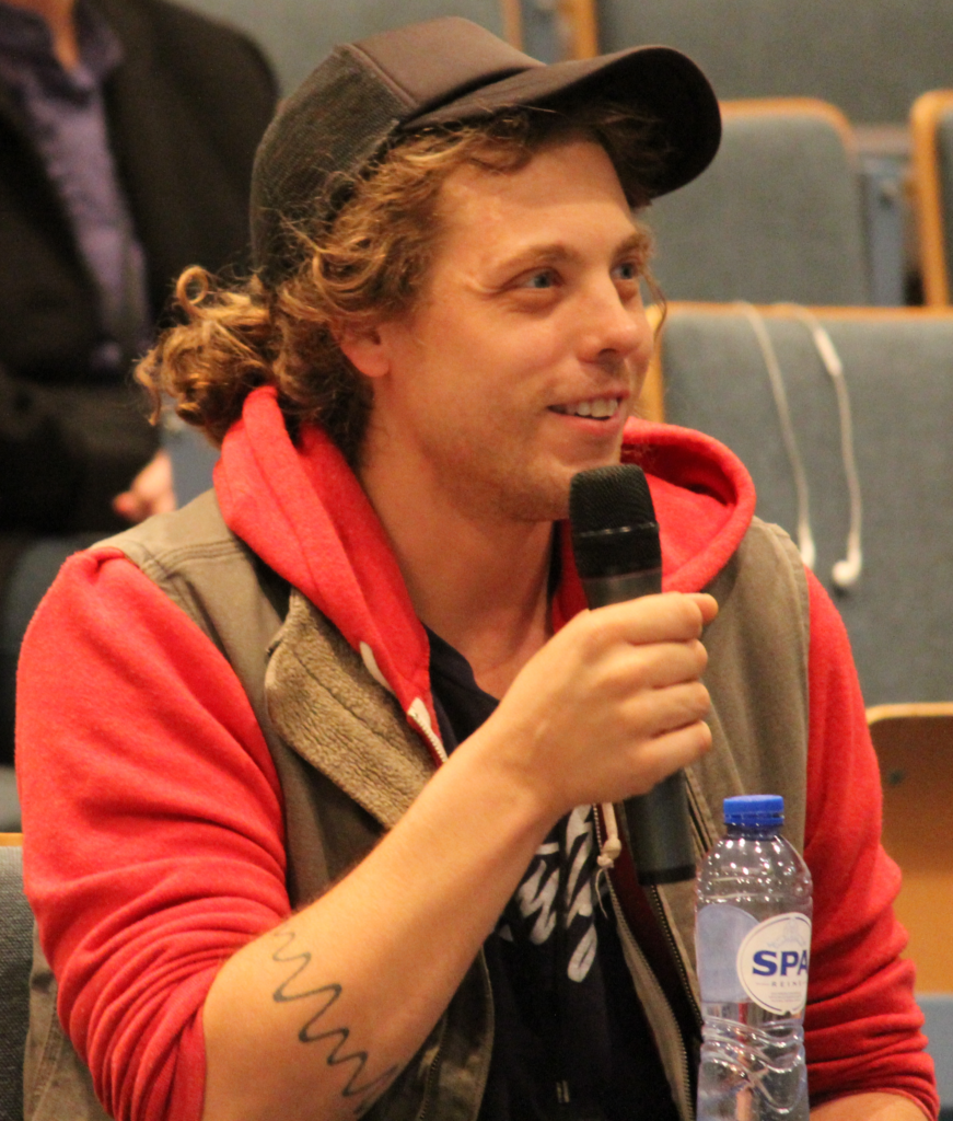 A man with light brown curly hair wearing a cap and orange hoodie, smiling and holding a microphone, posing a question after a conference presentation